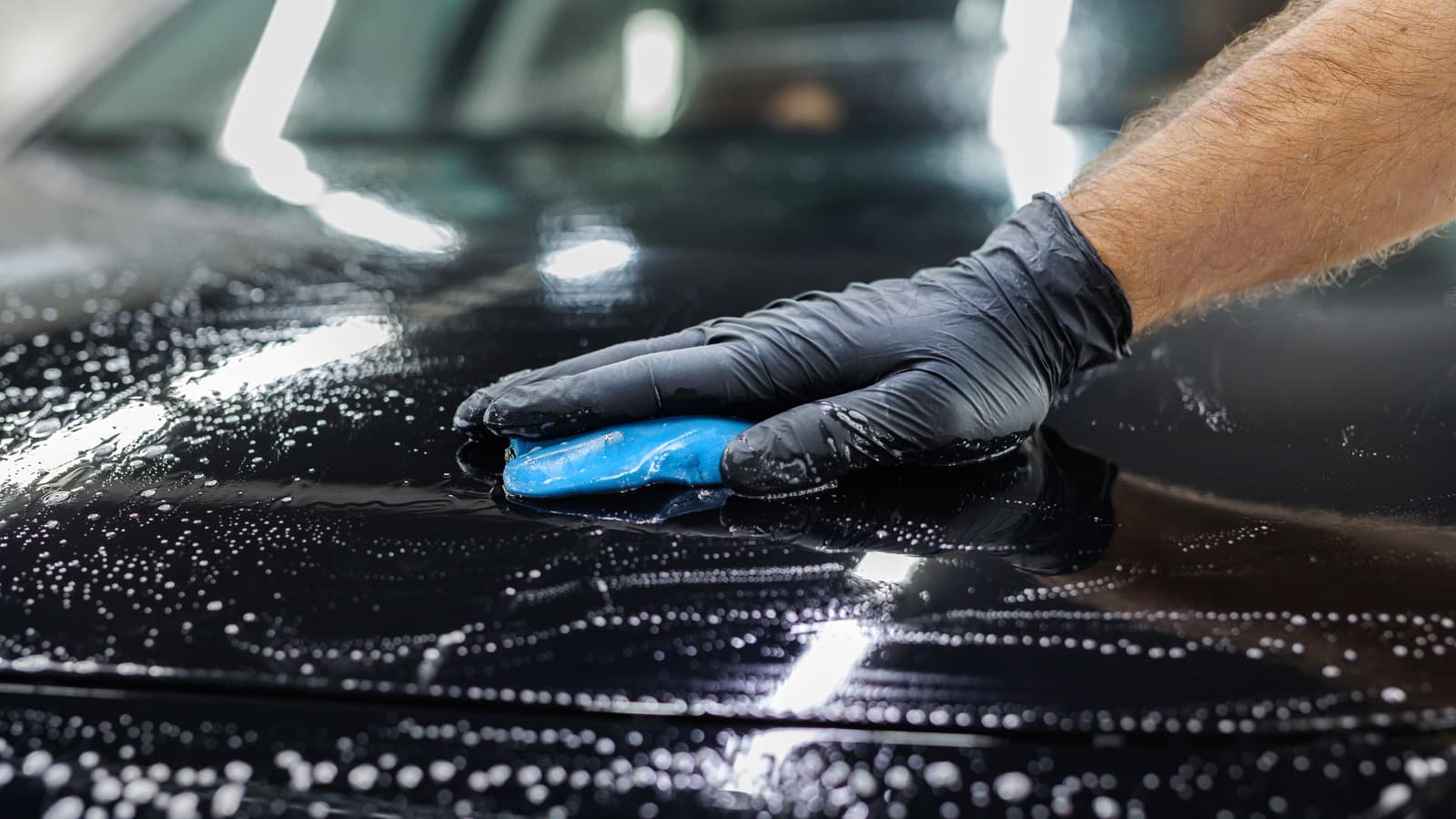 Gloved hand passing a clay bar across the wet, sudsy hood of a black car